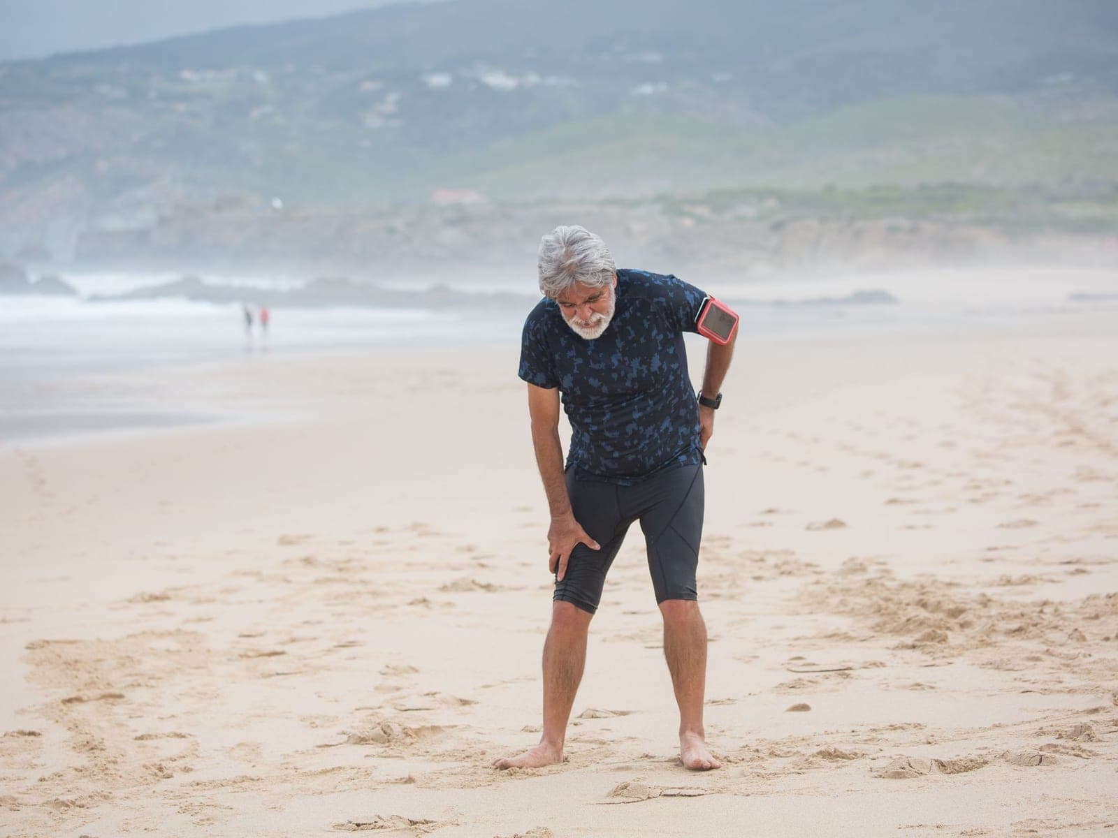 An older man pausing on a beach walk to hold his knee