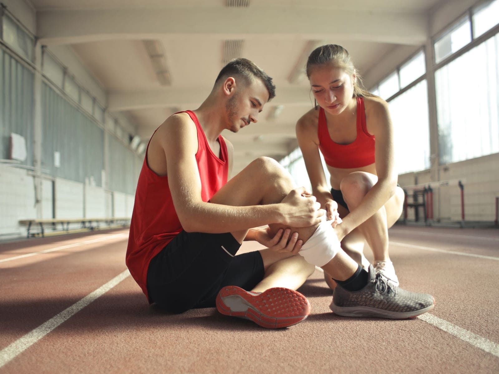 An athlete on an indoor track holding his ankle while a teammate checks it