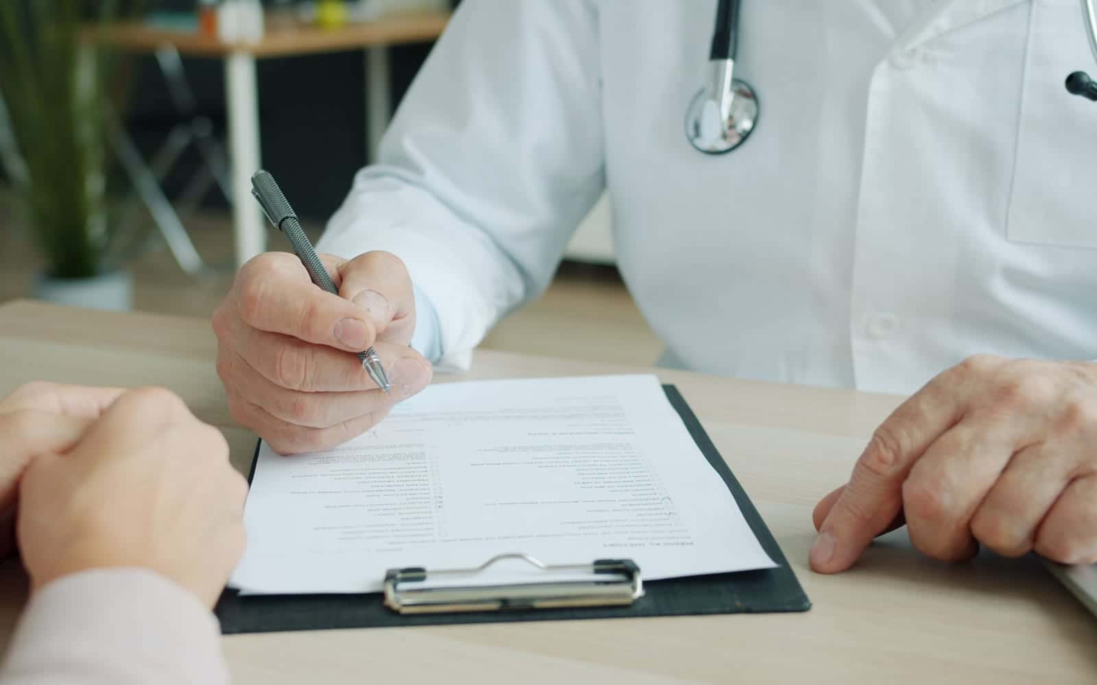 A doctor filling in a referral form on a clipboard across the desk from a patient